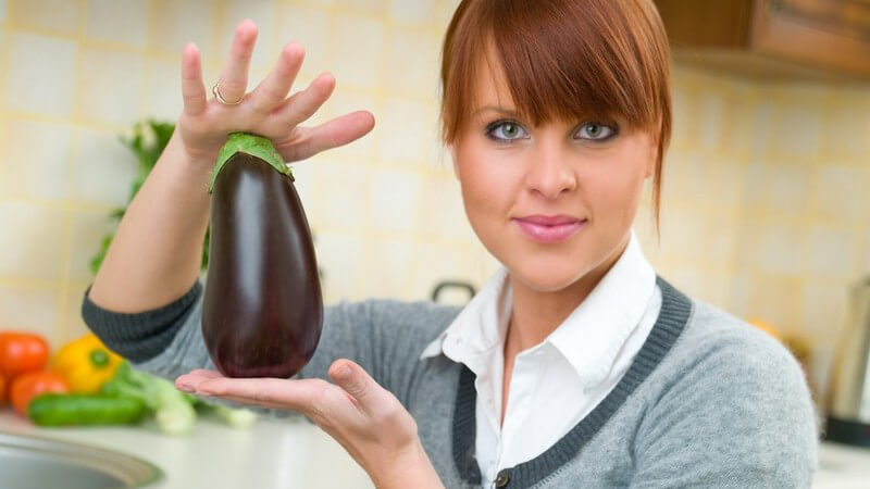 Junge Frau in Küche macht Salat und hält frische Aubergine in Kamera