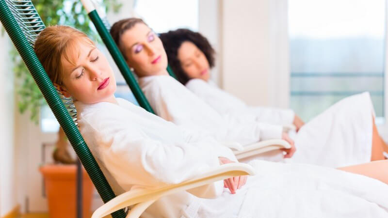 Drei Frauen in weißen Bademänteln entspannen im Liegestuhl in einem Ruheraum im Wellnesshotel