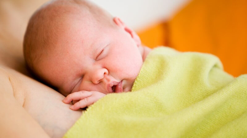 Schlafendes Baby mit leicht geöffnetem Mund liegt an Brust der Mutter unter hellgrüner Fleecedecke