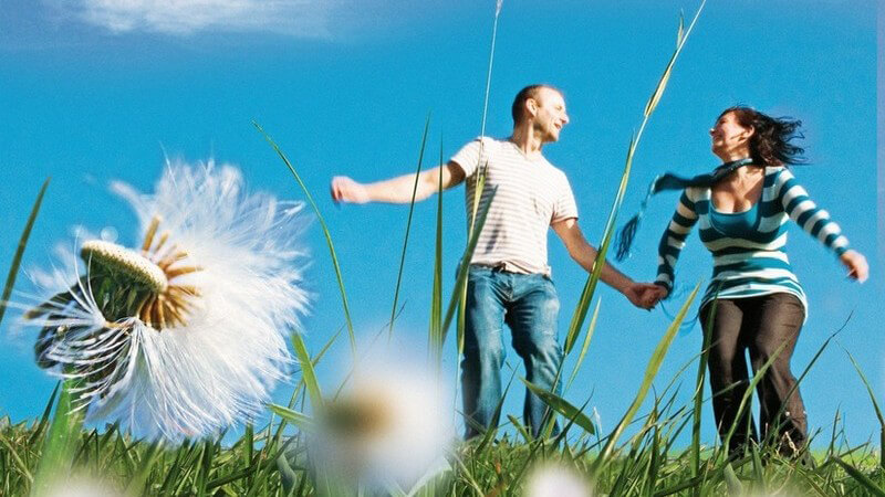 Pärchen (junger Mann in T-Shirt und junge Frau in gestreift) auf Wiese mit Blumen, blauer Himmel, halten Hände