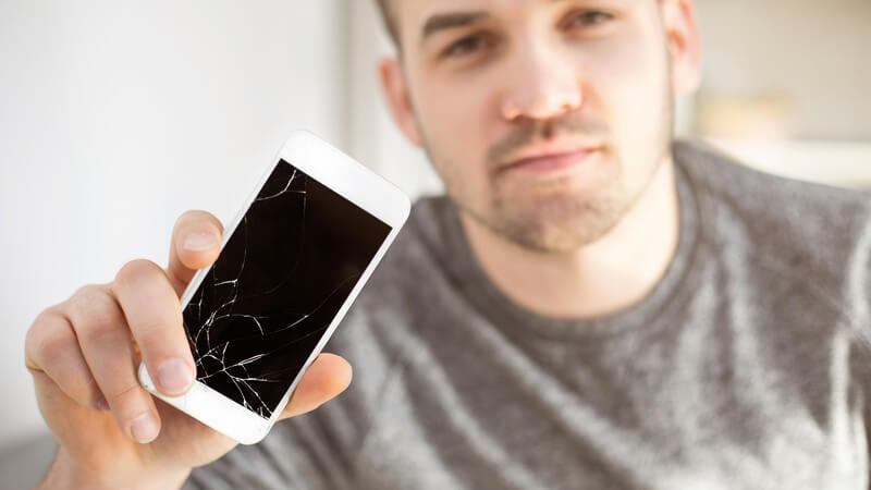 Junger Mann zeigt sein weißes Smartphone mit kaputter Glasoberfläche