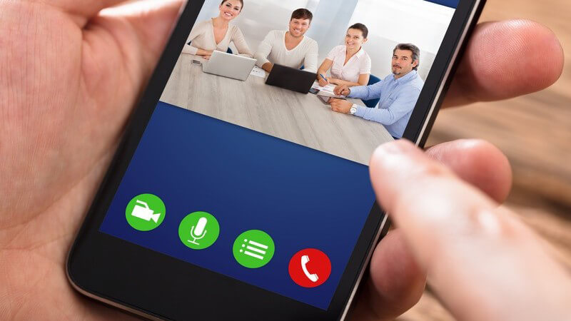 Smartphone zeigt eine Videokonferenz mit vier Geschäftsleuten am Tisch