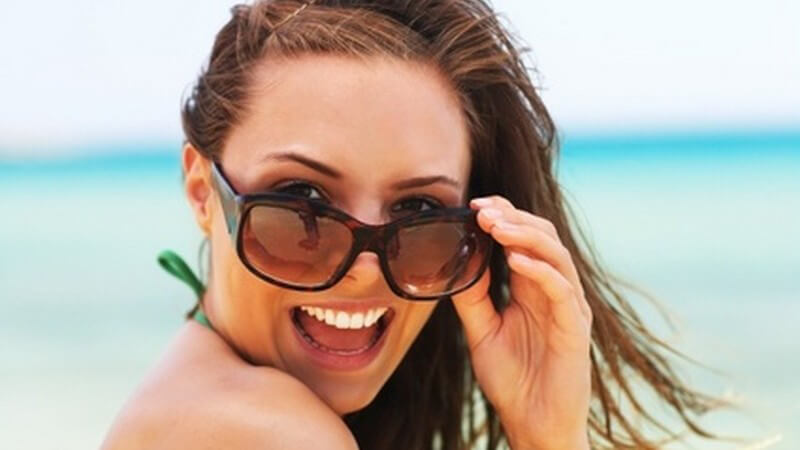 Gesicht einer jungen, braunhaarigen Frau in Bikini am Strand mit Sonnenbrille und offenem Mund