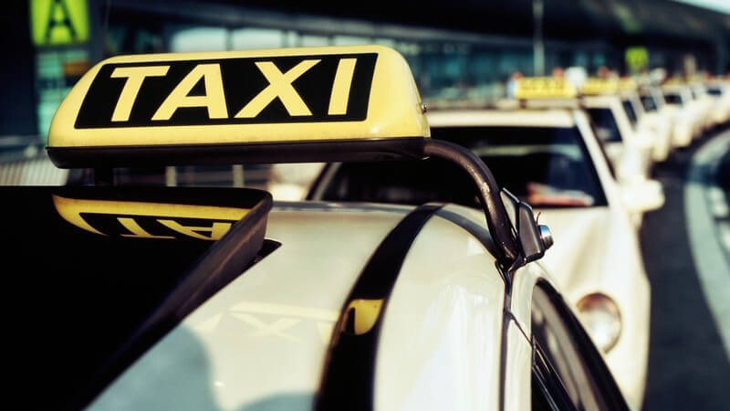 Nahaufnahme Schild eines Taxis, im Hintergrund lange Reihe aus Taxis am Flughafen