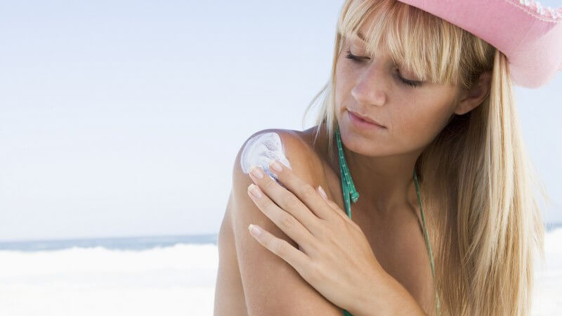Junge blonde Frau in Bikini mit Strandhut am Strand vor Meer trägt sich Sonnencreme o Sonnenschutz auf Arm auf