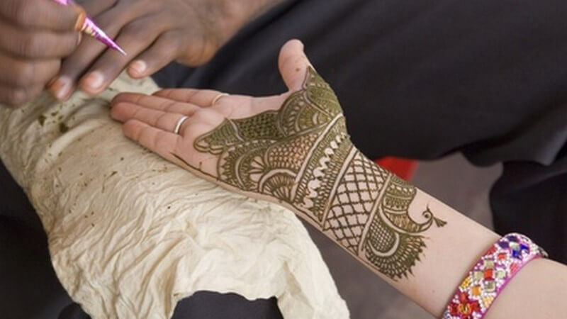 Hand einer Frau liegt auf Knie eines Mannes, exotische Henna-Bemalung, Mann verziert mit Spritze das Henna-Tattoo