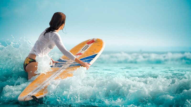 Junge Frau im Bikini und weißem, langarmigen Top steigt mit Surfbrett in die Wellen