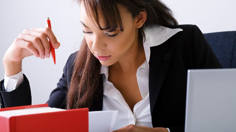 Beruf - Junge Frau in Bluse und Blazer sitzt mit einem roten Kulli konzentriert vor einem roten Buch