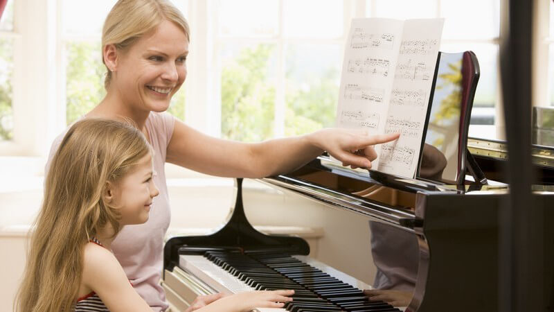 Kleines Mädchen sitzt am Klavier und spielt nach Noten, daneben sitzt Mutter, beide lächeln