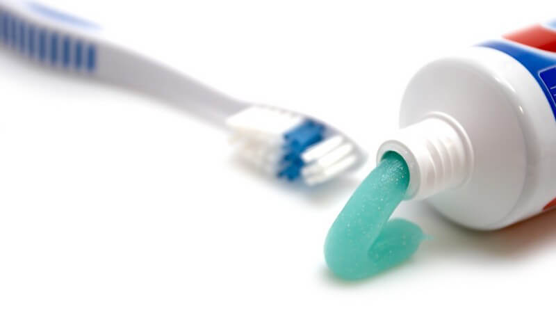 Links im Hintergrund weiß-blaue Zahnbürste, rechts offene Tube, aus der türkise Zahnpasta kommt
