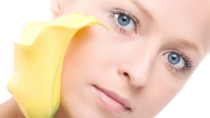 Gesicht einer jungen Frau mit blauen Augen, sie hält gelbe Blume und schaut in Kamera