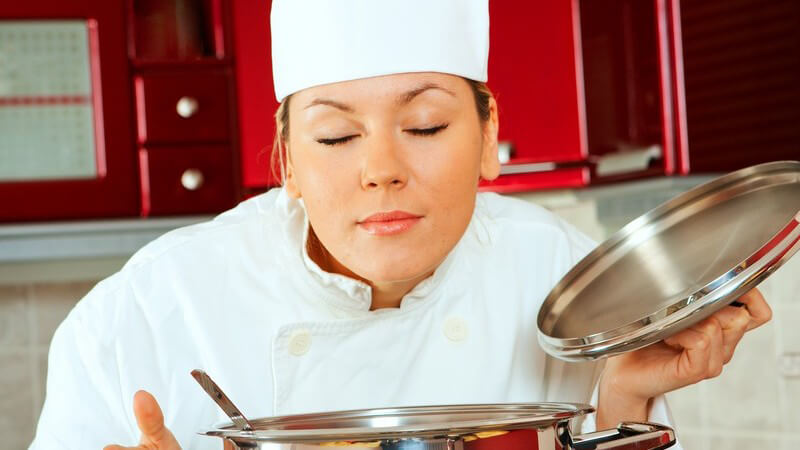 Junge Köchin mit geschlossenen Augen über Kochtopf gebeugt riecht genüsslich an Essen