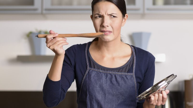 Frau in blauer Kochschürze steht am Topf, probiert von einem Holzlöffel das Essen und verzieht das Gesicht