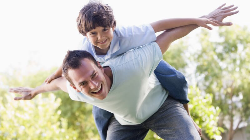 Junger Vater trägt seinen Sohn auf dem Rücken und spielt mit ihm Flieger