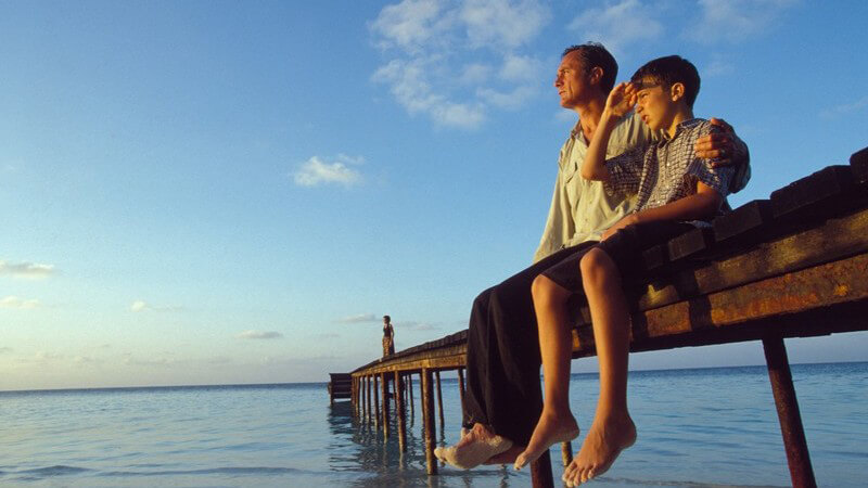 Vater und Sohn sitzen auf Bootssteg am Meer, schauen in den Sonnenuntergang, blauer Himmel