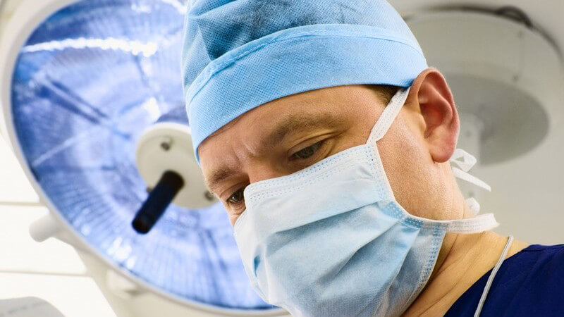 Gesicht eines Chirurgen mit Mundschutz, darüber Lampe in Operationssaal