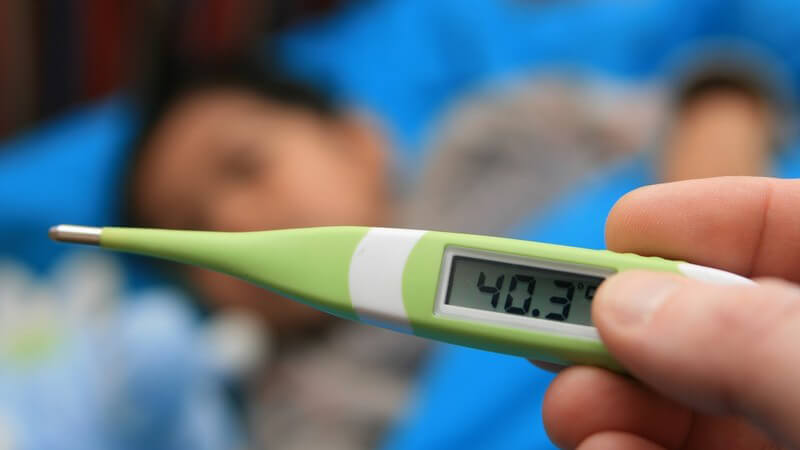 Fieberthermometer in Hand zeigt 40,3 Grad, im Hintergrund krankes Kind im Bett
