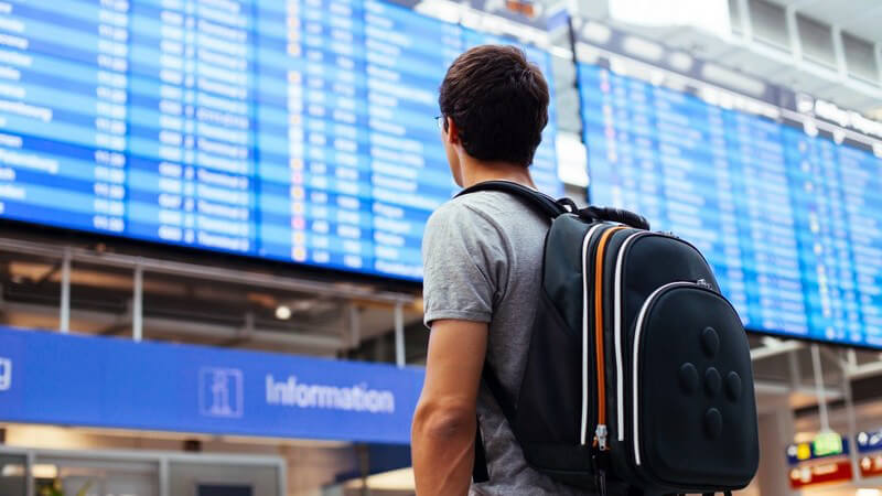 Mann mit Rucksack am Flughafen steht vor Anzeigetafel mit Abflug- oder Landezeiten der Flugzeuge
