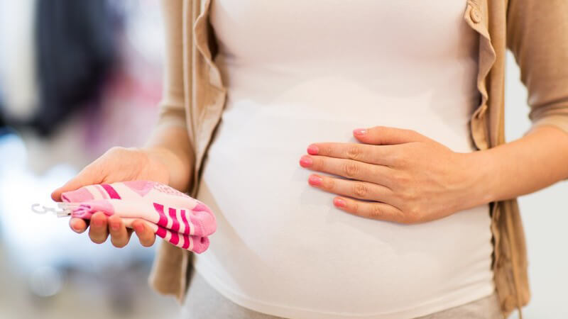 Schwangere Frau im Geschäft hält ein paar pinke Babysocken und eine Hand auf ihrem Babybauch