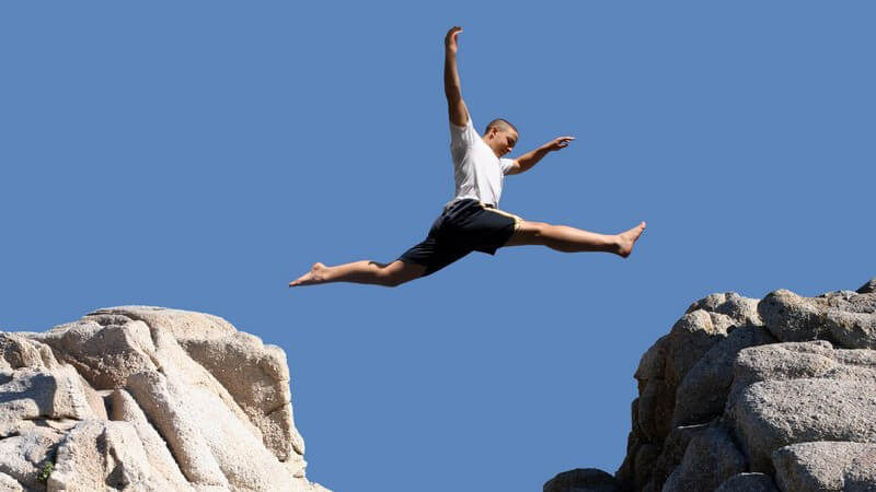 Mann in schwarzer Shorts und weißem Shirt springt von einem Felsen zum anderen, befindet sich gerade in der Luft