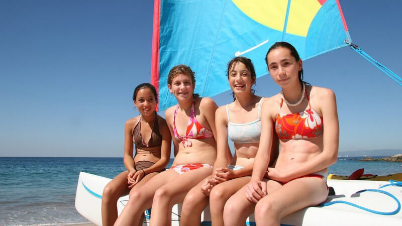 4 junge Mädchen im Bikini sitzen auf einem Segelboot mit blauem Segel
