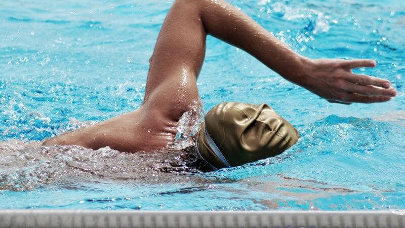 Schwimmer bei Kraulen im Schwimmbad mit goldener Badekappe, Gesicht im Wasser, linker Arm oben