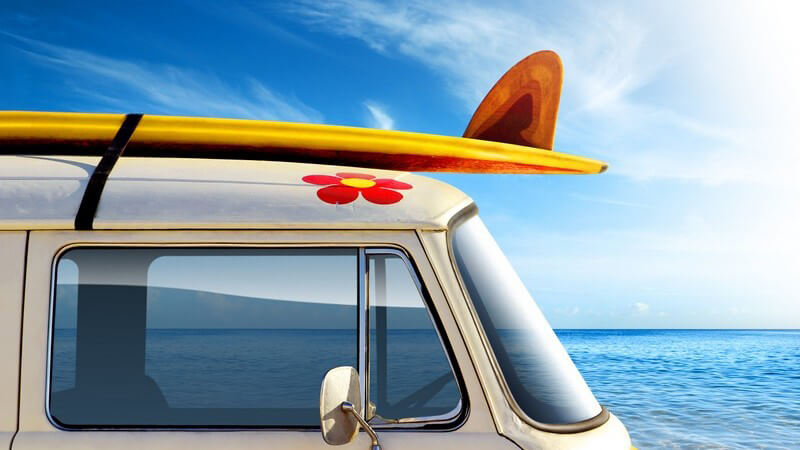 Van mit gelbem Surfbrett auf dem Dach, im Hintergrund Meer und blauer Himmel
