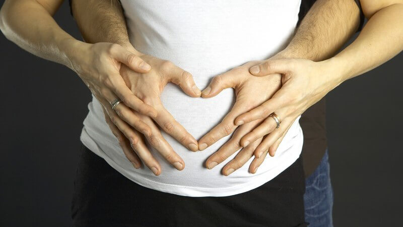 Bauch einer Schwangeren, von hinten von Männerhänden gehalten, die Herz formen