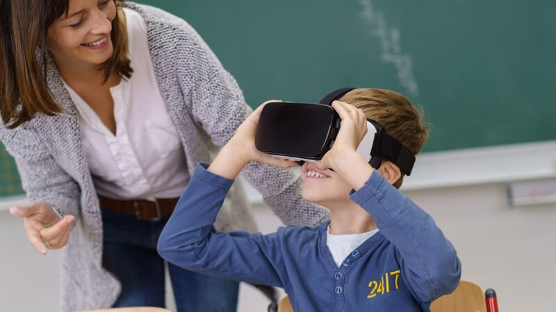 Junge in blauem Pullover sitzt im Klassenzimmer und guckt durch eine Virtual Reality-Brille, die Lehrerin steht daneben
