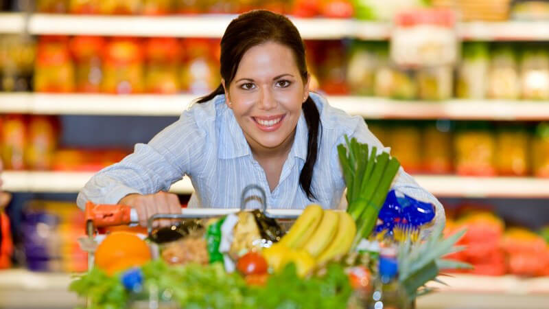 Junge lächelnde Frau schiebt vollen Einkaufswagen durch Supermarkt