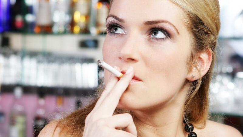 Junge Frau in schwarzem Top steht an Bar und zieht an Zigarette, schaut nach oben