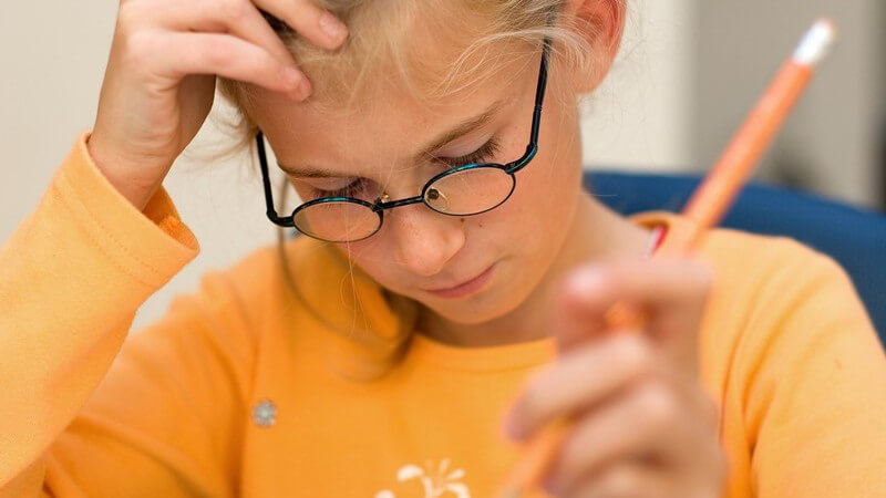 Mädchen mit Brille, Stift und orangem Pullover grübelt über einer Aufgabe