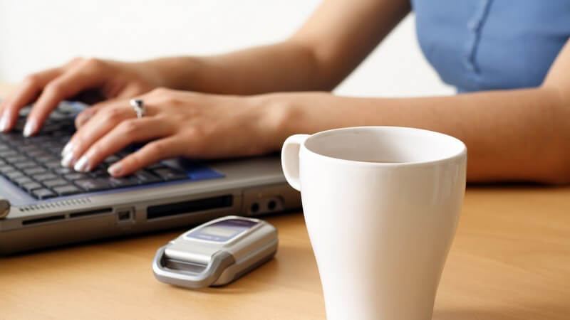 Hände einer Frau auf Notebook Tastatur, daneben Becher Kaffee und Handy