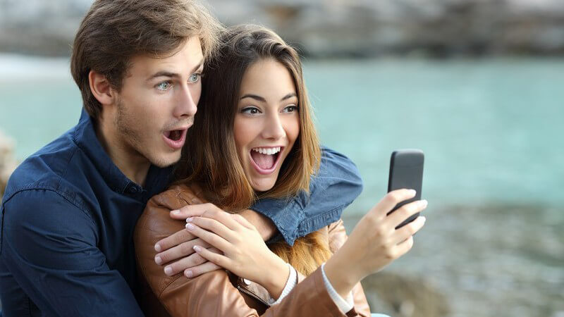 Junges Paar sitzt zusammen draußen und guckt schockiert auf ein Smartphone