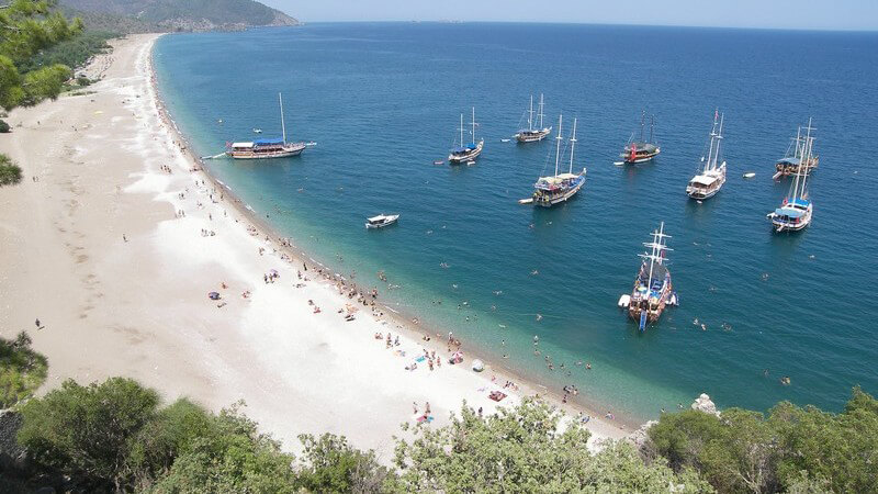 Boote auf blau-türkisem Meer vor weißem Strand, im Hintergrund Berge