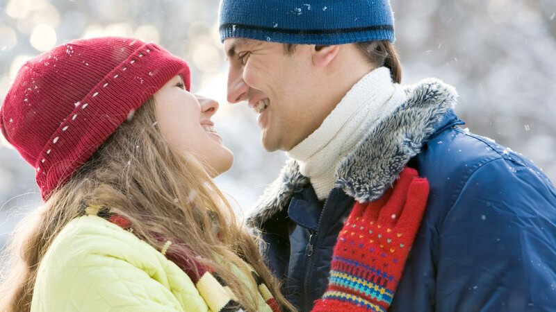 Junges Paar, warm angezogen steht draußen im Schnee und schaut sich verliebt an