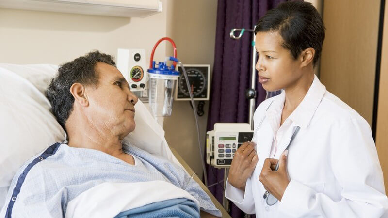 Junge Ärztin in weißem Kittel spricht mit älterem Patient, der im Krankenbett liegt