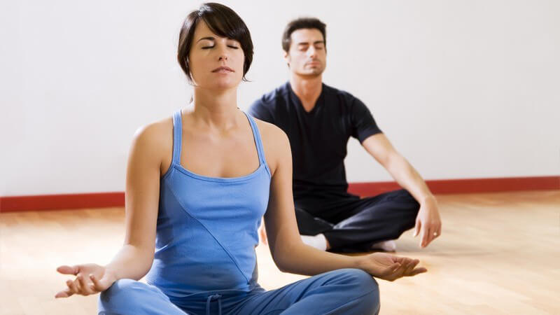 Holzboden, Frau in blauer Sportkleidung vorne, Mann mit schwarzer Sportkleidung hinten, Schneidersitz, Meditation