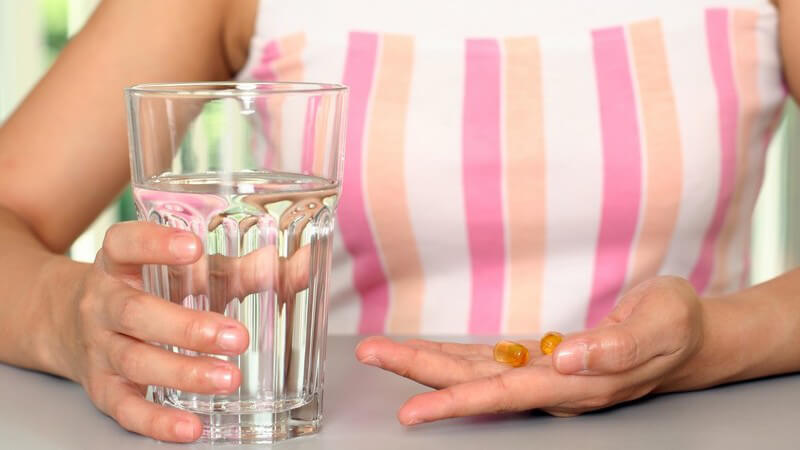 Frau sitzt mit einem Glas Wasser und Vitamintabletten in der linken Hand am Tisch