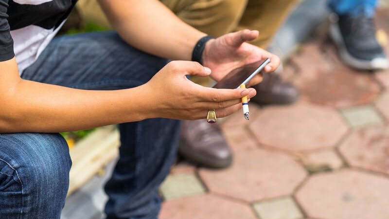 Junger Mann sitzt draußen, raucht eine Zigarette und beschäftigt sich mit seinem Smartphone