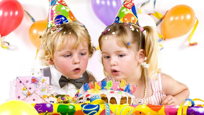 Kleiner Junge und Mädchen mit Partyhütchen auf Kindergeburtstag, schauen auf Kuchen, im Hintergrund bunte Luftballons
