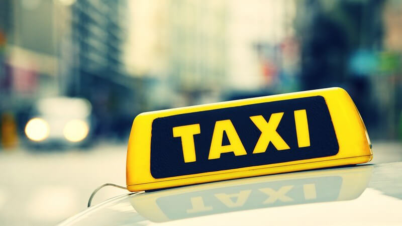 Gelbes Taxi-Schild auf dem Dach eines Taxis, verschwommen im Hintergrund Straßen und Hochhäuser