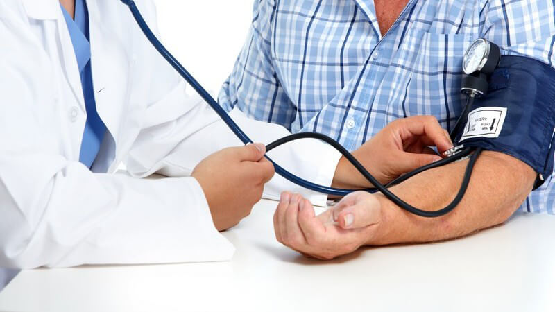 Arzt misst den Blutdruck am linken Arm eines Patienten in blauem Karohemd, hört ihn außerdem am Arm ab