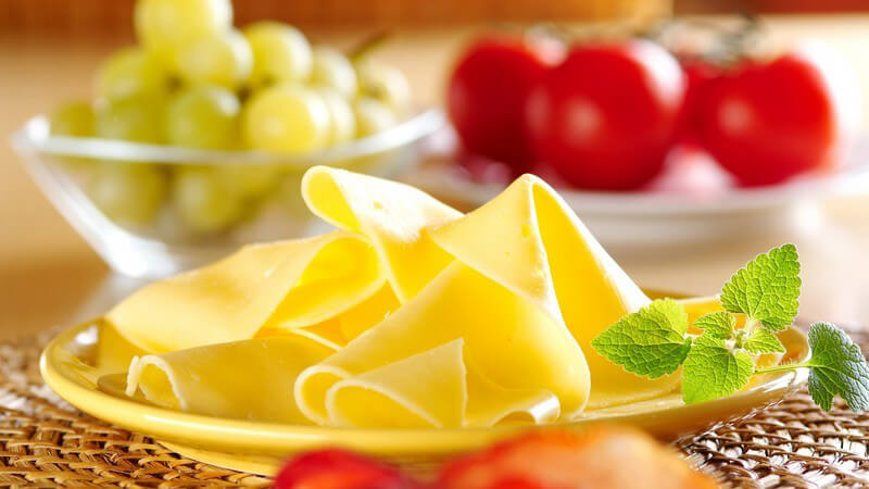 Gelbe Käsescheiben auf Teller, daneben frische Tomaten und Trauben auf Frühstückstisch