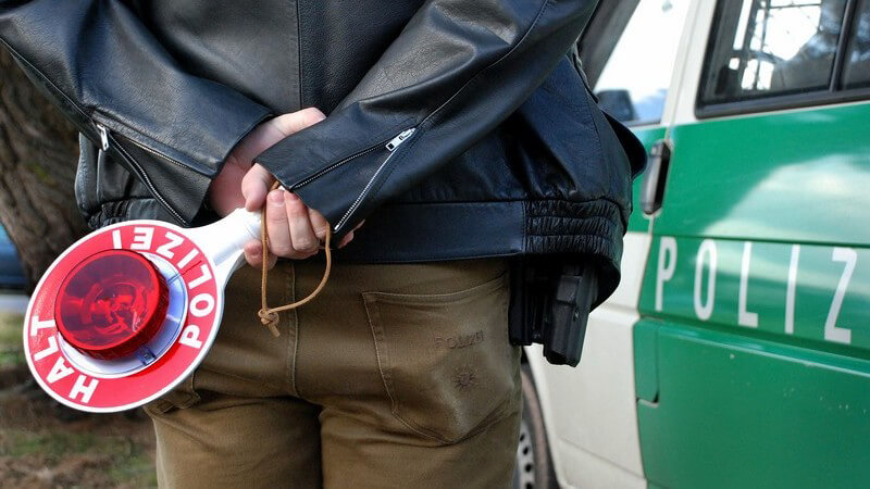 Rückenansicht Polizist, hält Kelle hinter Rücken vor einem Polizeiwagen