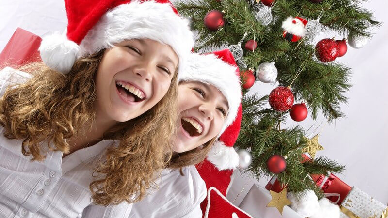 Zwei Mädchen mit Nikolausmütze sitzen vor Weihnachtsbaum, lachen