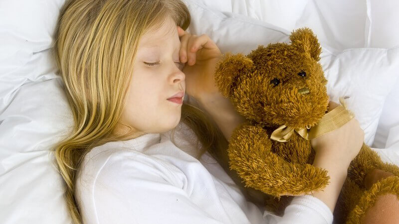 Blondes Mädchen liegt auf der Seite, hält ihren Teddy im Arm und schläft