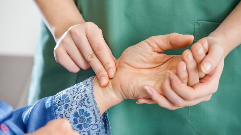 Krankenschwester in grünem Kittel überprüft am Handgelenk einer Seniorin den Puls