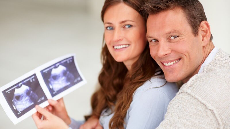 Paar hält ein Ultraschallbild von ihrem Baby, beide lächeln in Kamera