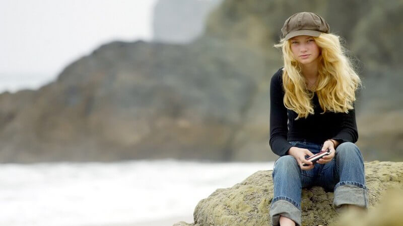 Mädchen mit blonden, langen Haaren und grüner Mütze vor Bergen und See